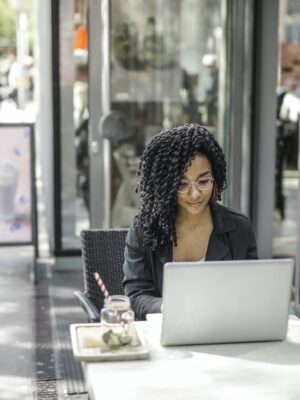 High angle of pensive African American female freelancer in glasses and casual clothes focusing on screen and interacting with netbook while sitting at table with glass of yummy drink on cafe terrace in sunny day