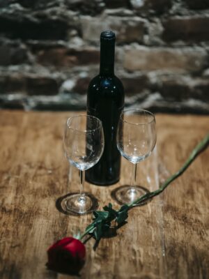 Wooden table with bottle of alcohol drink near wineglasses and red rose near brick wall in restaurant