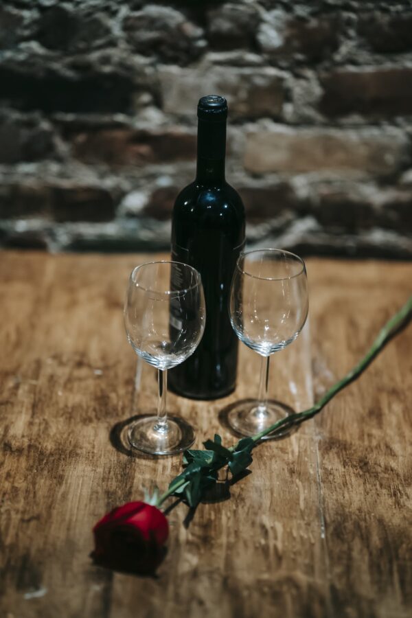 Wooden table with bottle of alcohol drink near wineglasses and red rose near brick wall in restaurant